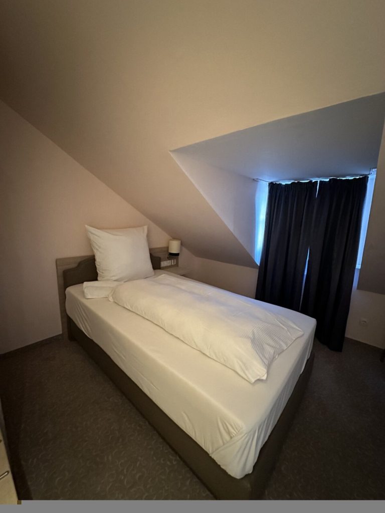 Einzelbett mit weißer Bettwäsche in einem Zimmer mit schräger Decke und dunklen Vorhängen am Fenster.